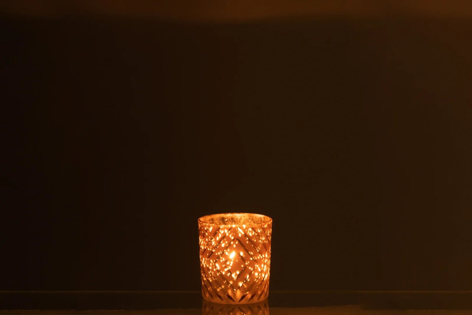 De Theelichthouder Zigzag Glas Zalm Klein werpt een warme, zachte gloed tegen een donkere achtergrond en creëert zo een gezellige en serene sfeer.