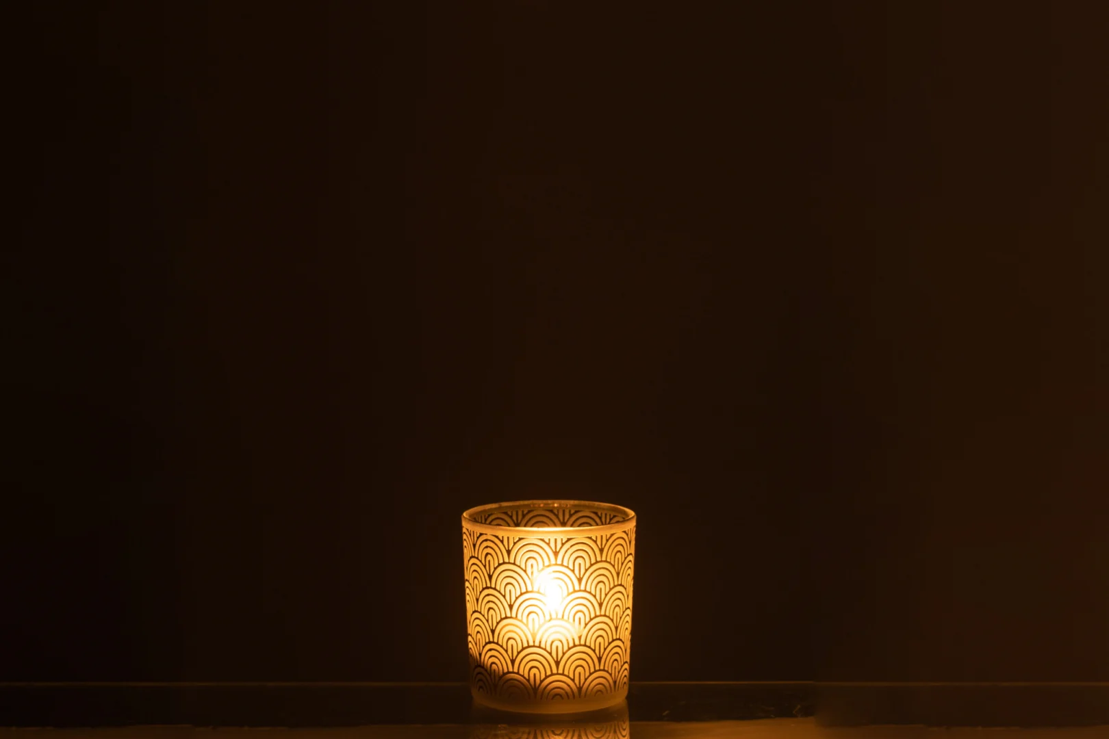 Een brandende kaars gloeit warm in de Theelichthouder Boog Glas Groen Klein, de decoratieve houder met sint-jakobsschelpmotief verlicht de donkere, lege ruimte met een zacht, uitnodigend licht.