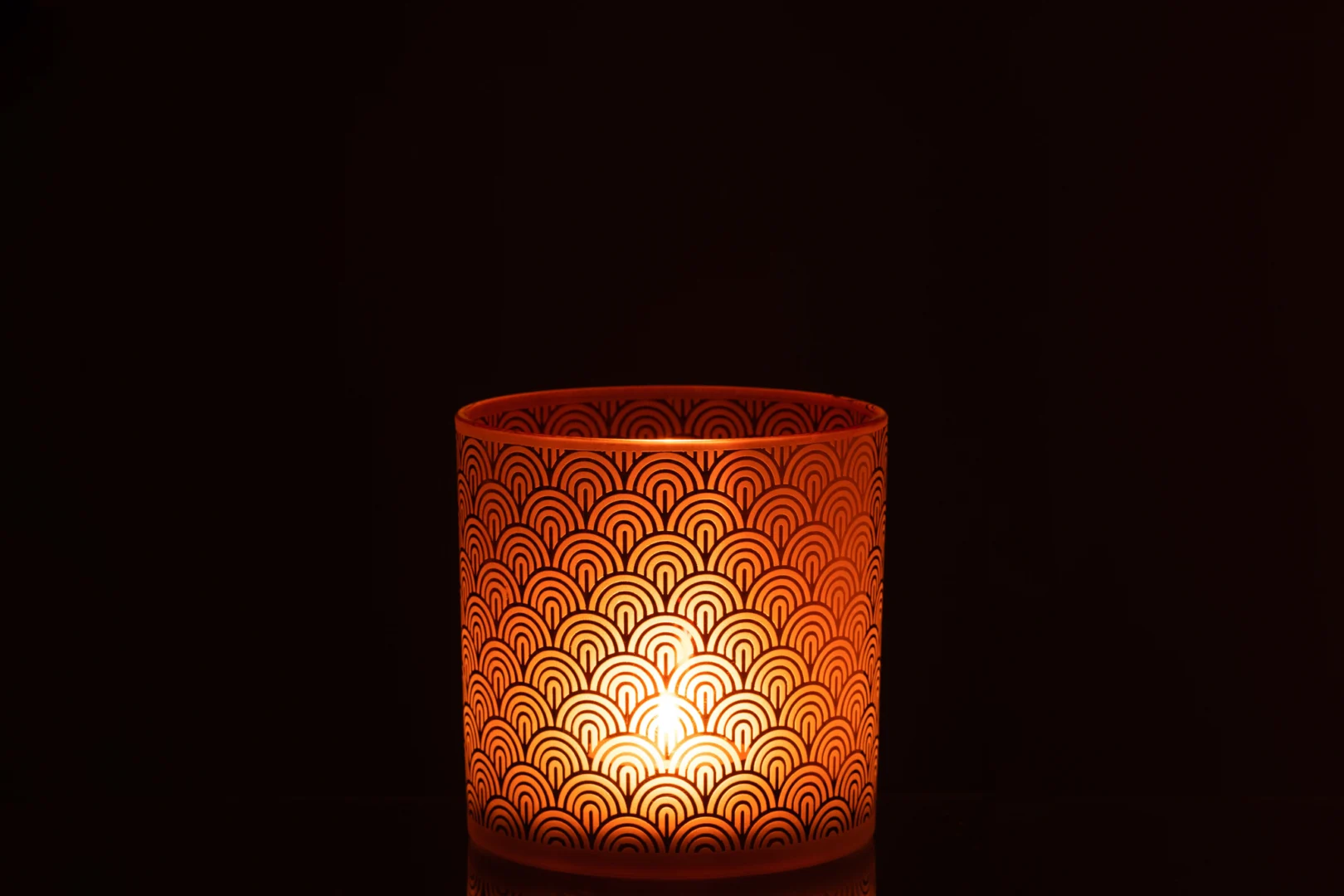 De Kaarsenhouder Orkaan Arc Glas Oranje Groot heeft een brandende kaars in de oranje glazen houder met sint-jakobsschelpmotief, die een warm licht werpt tegen een donkere achtergrond.