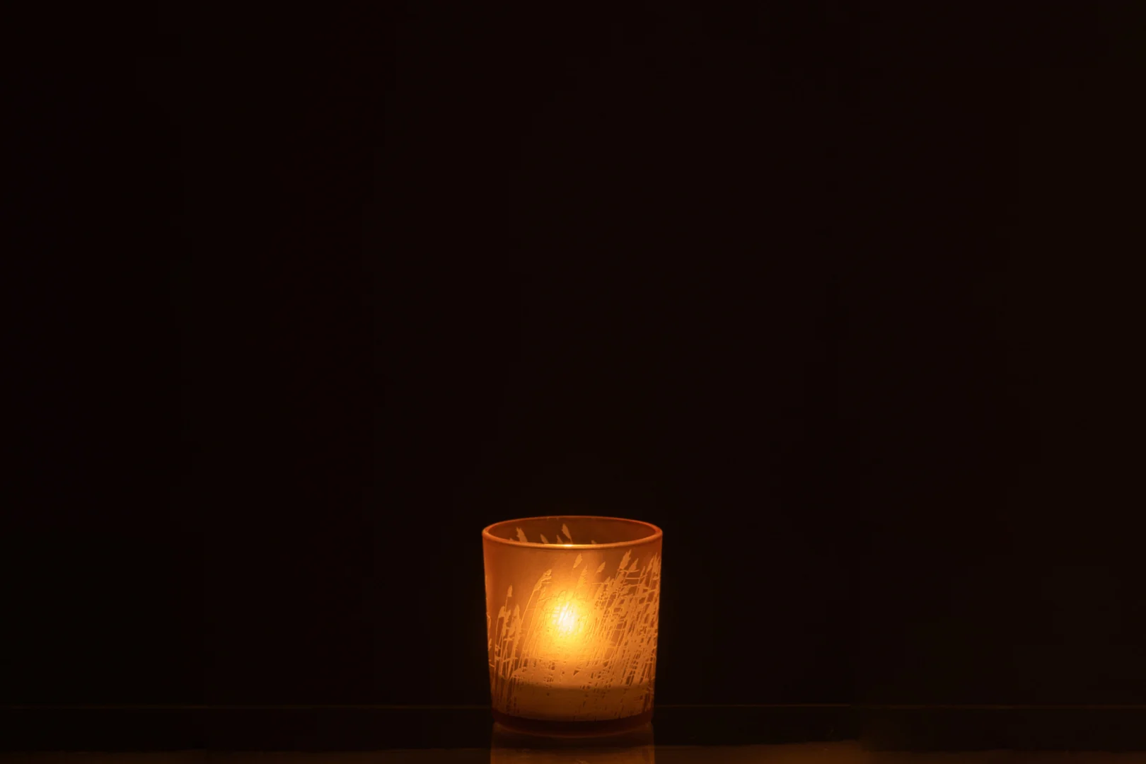 Een enkele Theelichthouder Gras Glas Beige Klein kandelaar rust op een donker oppervlak tegen een zwarte achtergrond en werpt een warme, zachte gloed door het matglas.