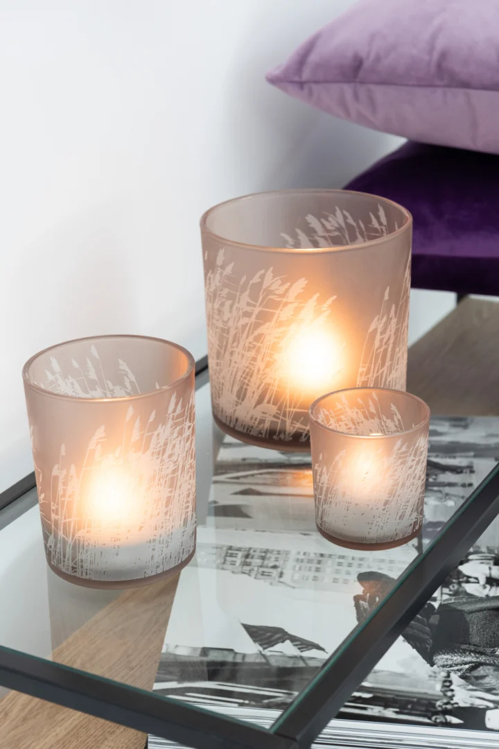 Drie Theelichthouder Gras Glas Beige Klein kandelaars met gloeiende kaarsen erin staan op een glazen tafel met een grasachtig patroon. Een foto en twee paarse kussens staan op de achtergrond.