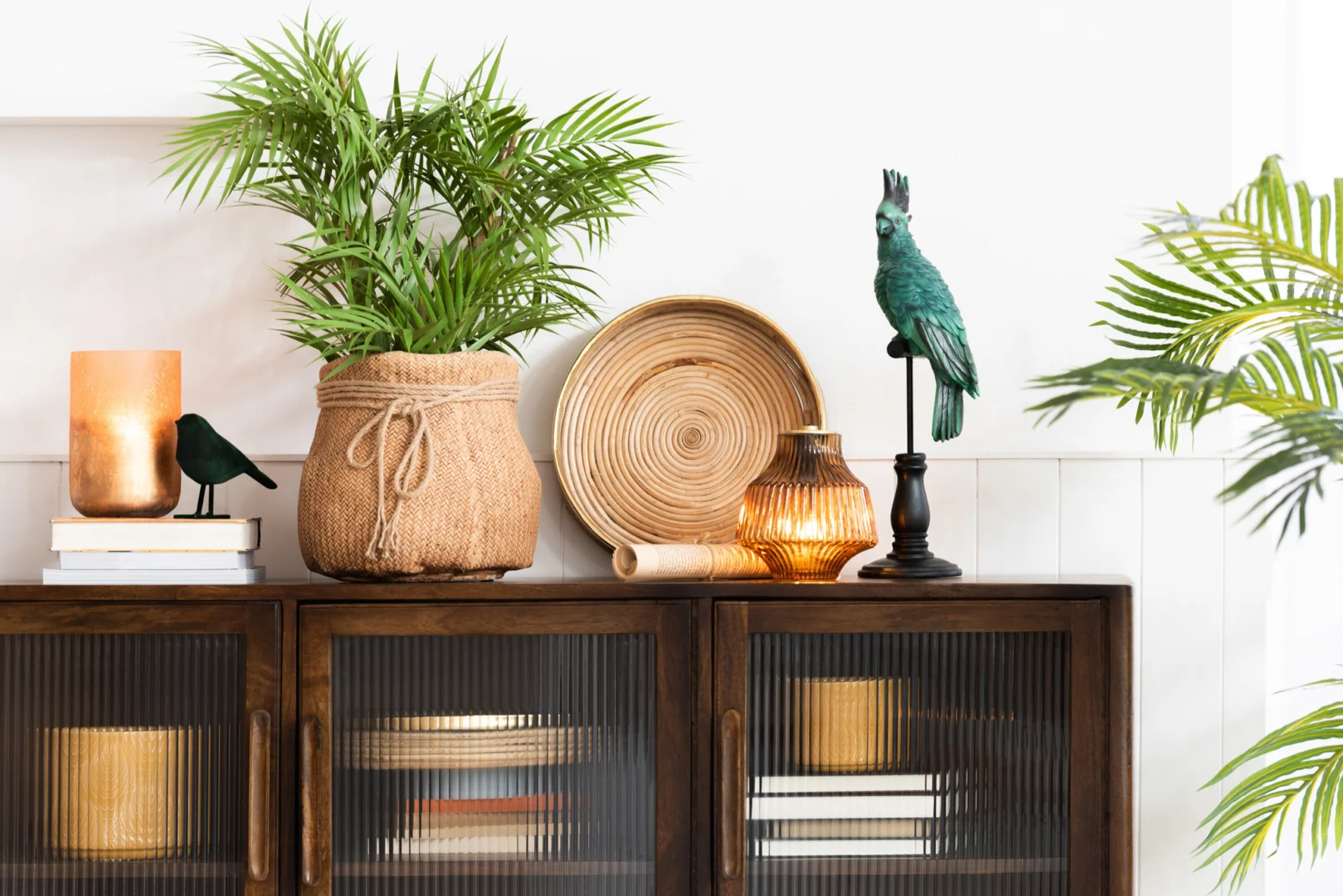 Een houten kast met daarop decor: een potplant in een gevlochten mand, een gevlochten bord, de Kaarsenhouder Orkaan Streep Glas Goud Oker Groot, een groenblauw vogelbeeldje, gestapelde boeken en rechts een bladplant.