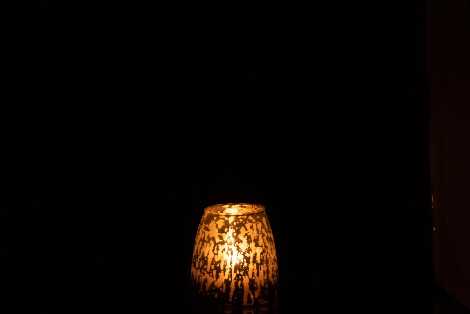 De Theelichthouder Melkglas Grijs Klein straalt een zachte gloed uit door het decoratieve grijze glas en verlicht de omgeving zacht tegen een donkere achtergrond.