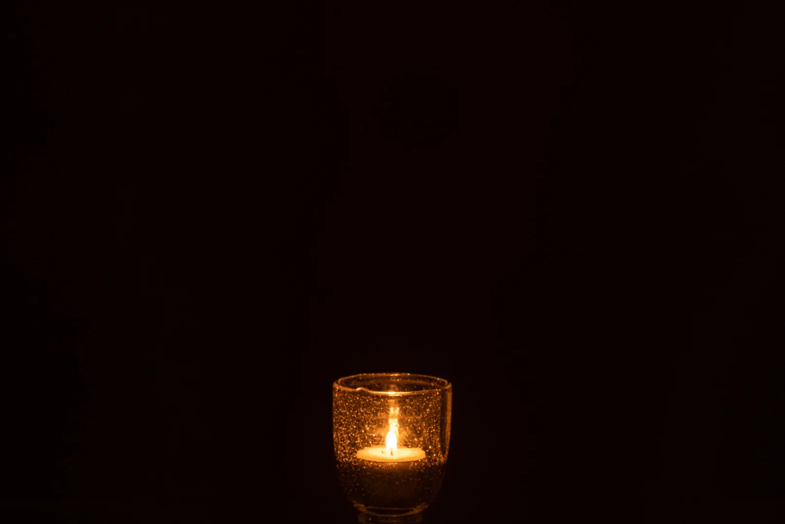 De Theelichthouder Bubbels Glas Transparant Klein houdt een gloeiende kaars vast, werpt een warm licht tegen een donkere achtergrond en creëert een serene, vredige sfeer.
