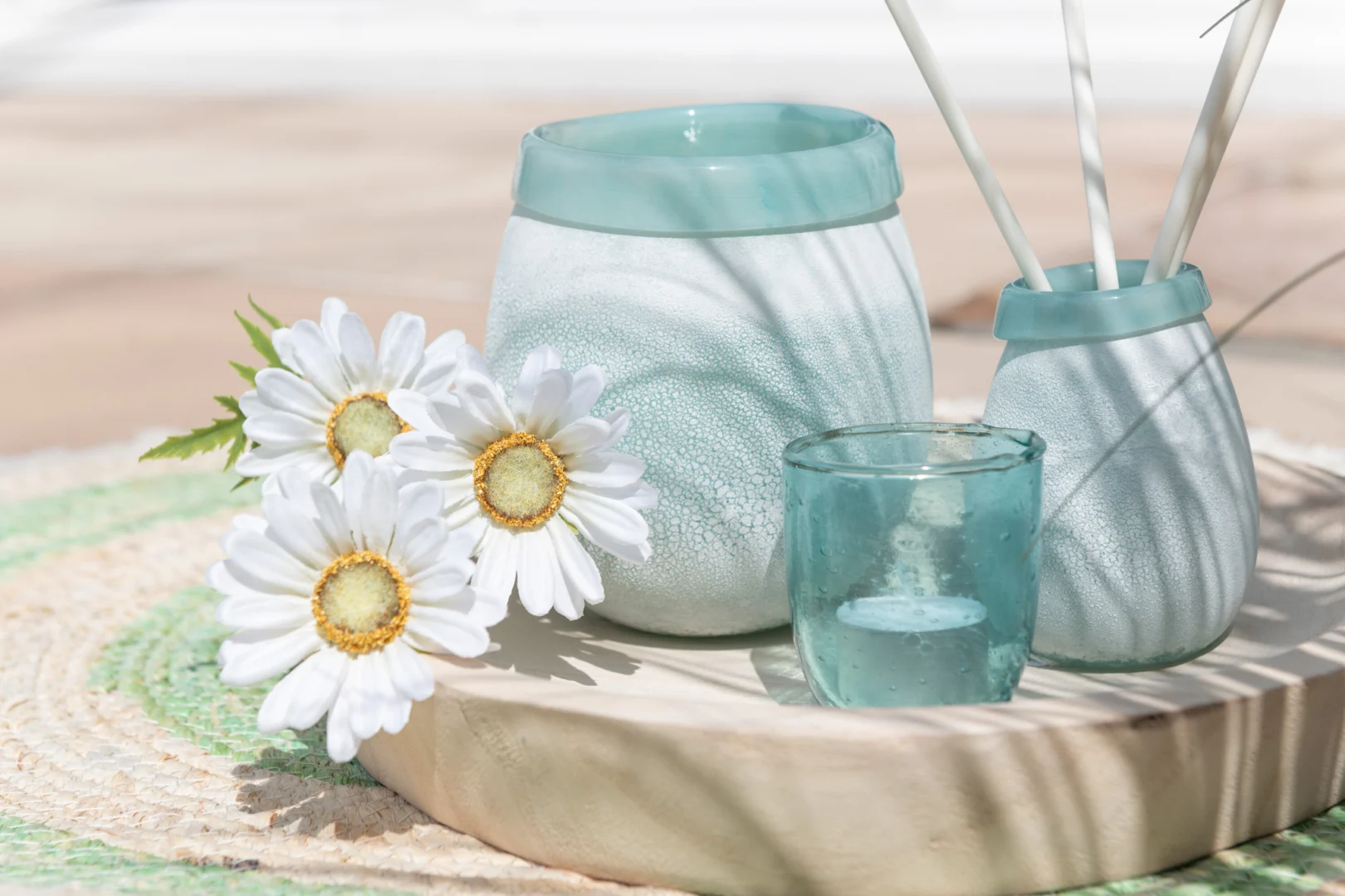 Drie witte madeliefjes staan naast blauwe matglazen potten en de Theelichthouder Bubbels Glas Aqua Klein op een rond houten dienblad, allemaal gerangschikt op een geweven mat in helder, natuurlijk zonlicht.