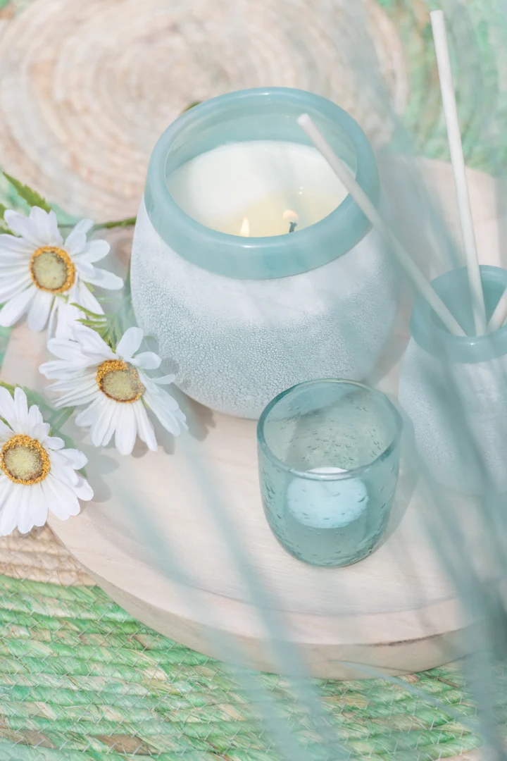 Een zacht, pastel tafereel met witte madeliefjes, een brandende kaars in een matblauwe houder, Theelichthouder Bubbels Glas Aqua Klein en een diffuserpotje op een licht houten dienblad boven een groene geweven mat.