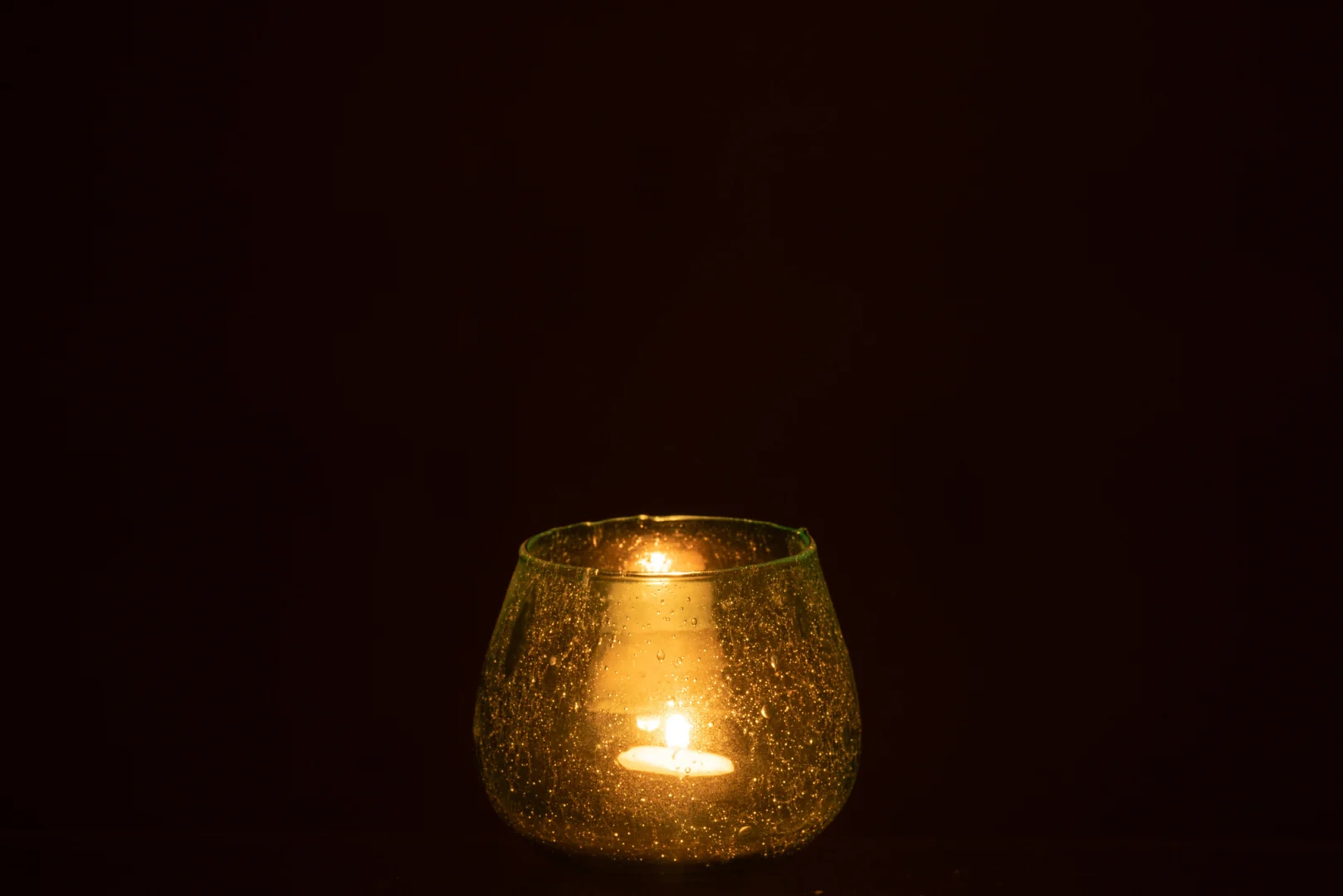 De Kaarsenhouder Orkaan Bellen Glas Aqua heeft een enkele brandende kaars die warm oplicht in een ronde, gespikkelde aquaglazen houder tegen een donkere achtergrond.