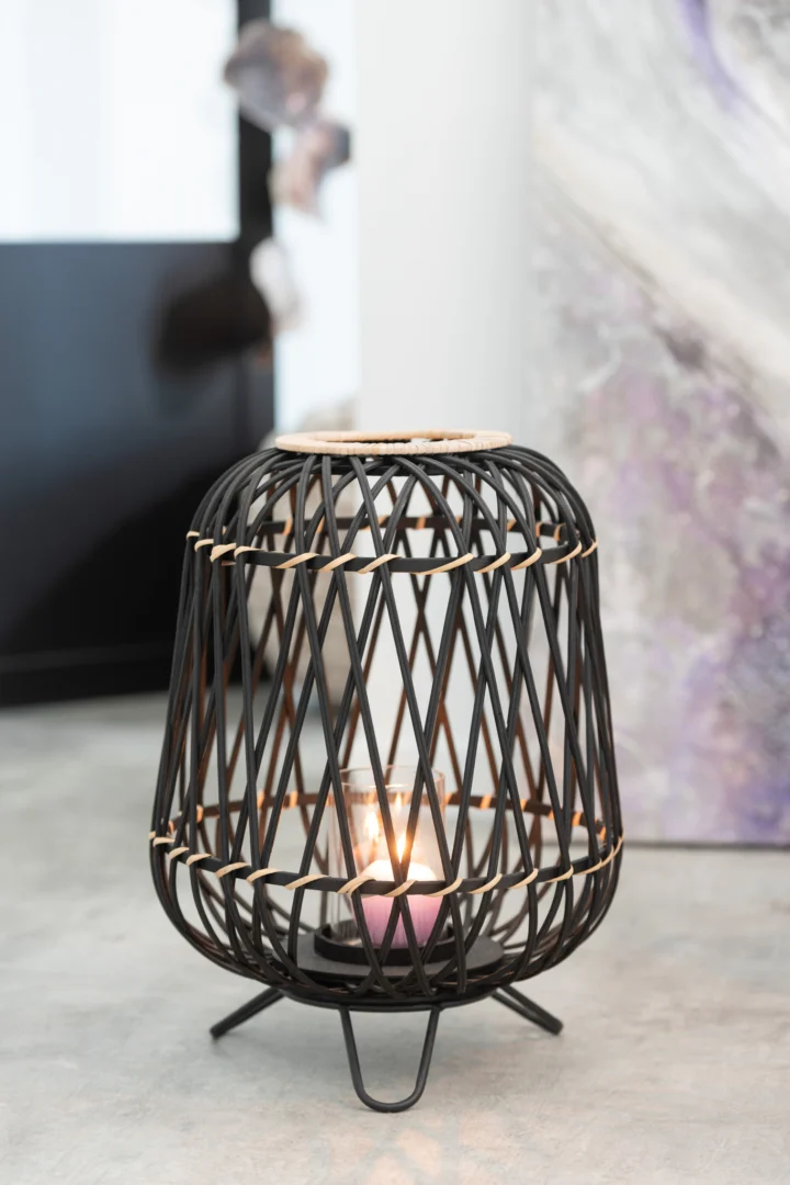 Een kleine kaars gloeit in de Kaarsenhouder Bamboe Zwart Natuurlijk Medium, een zwarte, opengewerkte bamboelantaarn met een ronde voet, die op een lichte vloer staat in de buurt van abstracte kunst en een muur.