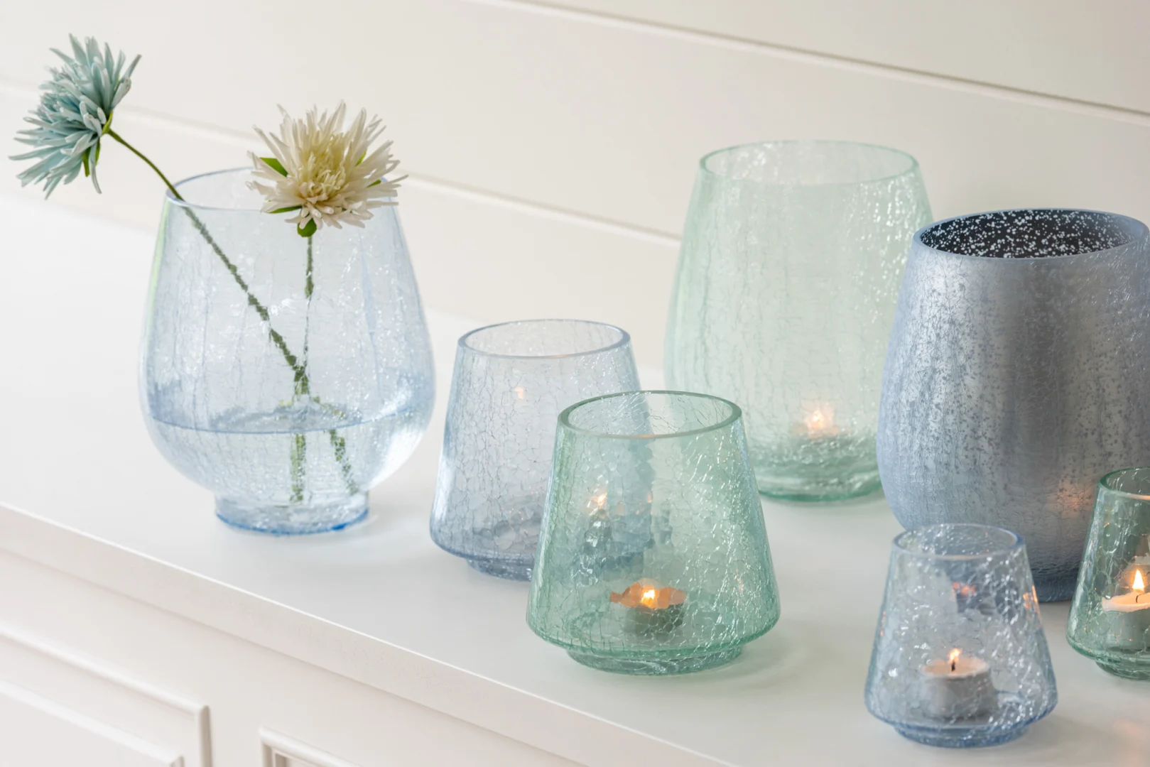 De Kaarsenhouder Orkaan Conic Craquelure Blauw Medium Set 2 heeft blauw glazen kandelaars met textuur, sommige met brandende waxinelichtjes, uitgestald op een wit oppervlak naast vazen met lichtblauwe en crèmekleurige bloemen.