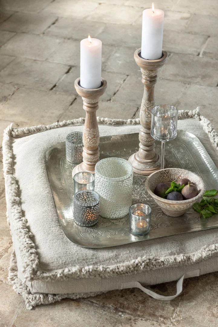De Kaarsenhouder Amoro Mangohout Wit Medium heeft witte pilaarkaarsen, gecombineerd met glazen votiefkaarsen, een schaal met vijgen en verse basilicum op een zilveren dienblad bovenop een crèmekleurig kussen met textuur op een stenen vloer.