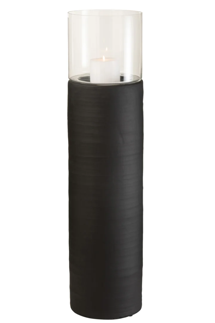 Een witte pilaarkaars wordt aangestoken in de Kaarsenhouder Orkaan Cement Glas Zwart-een helder glazen orkaanhouder met een hoge, matzwarte cilindrische voet.
