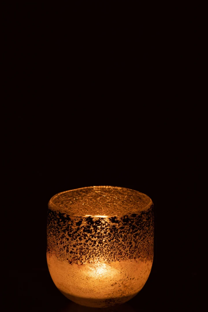 De Orkaan Milano Glas Grijs/Roze Klein is een kleine glazen kandelaar met textuur die een warme oranje gloed afgeeft en zacht licht werpt tegen een donkere achtergrond.