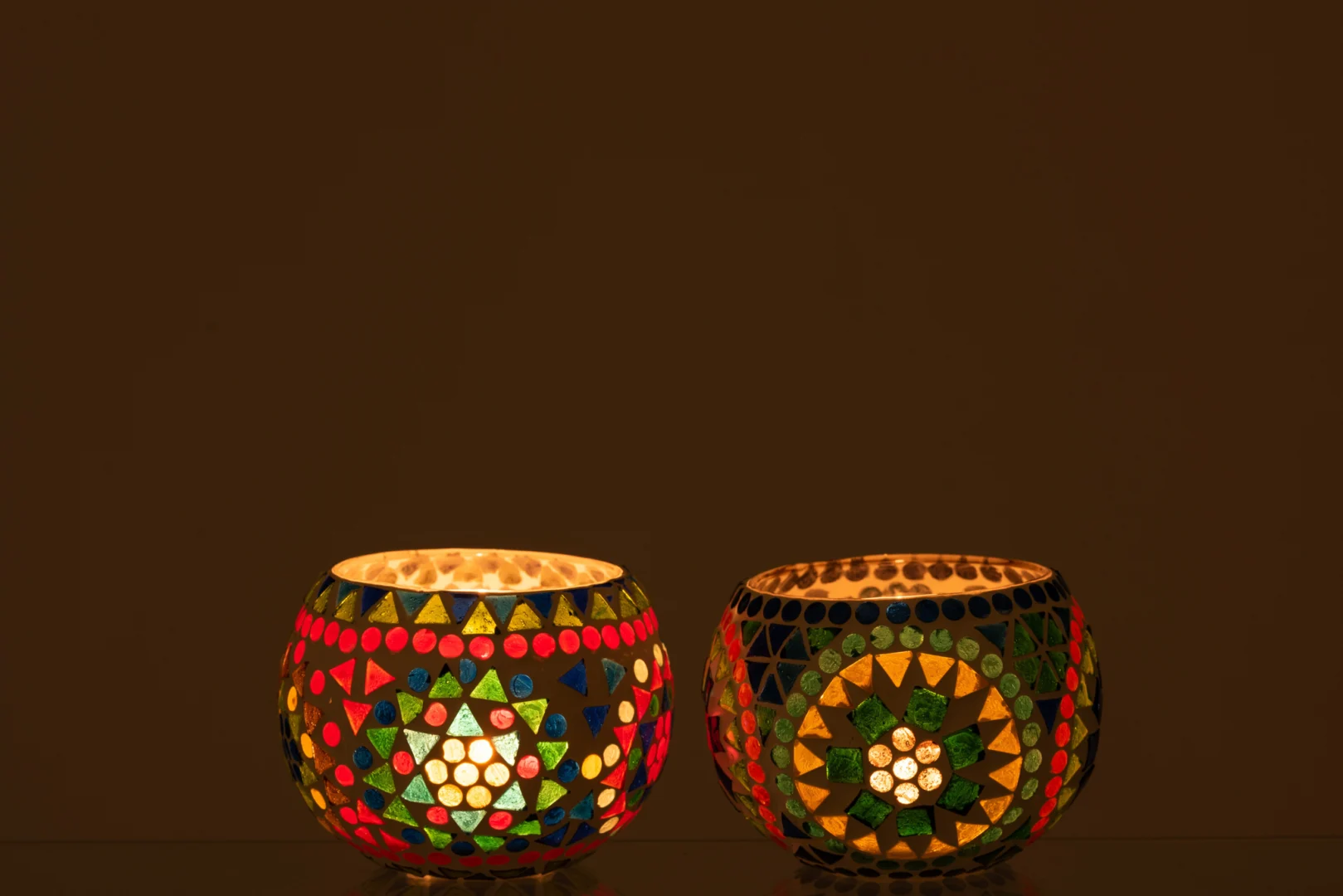 Theelichthouder Eki Mozaïek Glas Mix Medium Set 2 heeft twee kleurrijke mozaïek glazen kandelaars met brandende kaarsen erin, naast elkaar geplaatst tegen een donkere achtergrond, die een warme, uitnodigende gloed verspreiden.