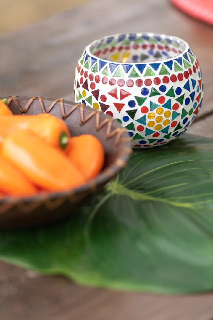De Theelichthouder Eki Mozaïek Glas Mix Medium Set 2 staat op een groot groen blad naast een houten schaal met kleine oranje paprika's op een houten tafel.
