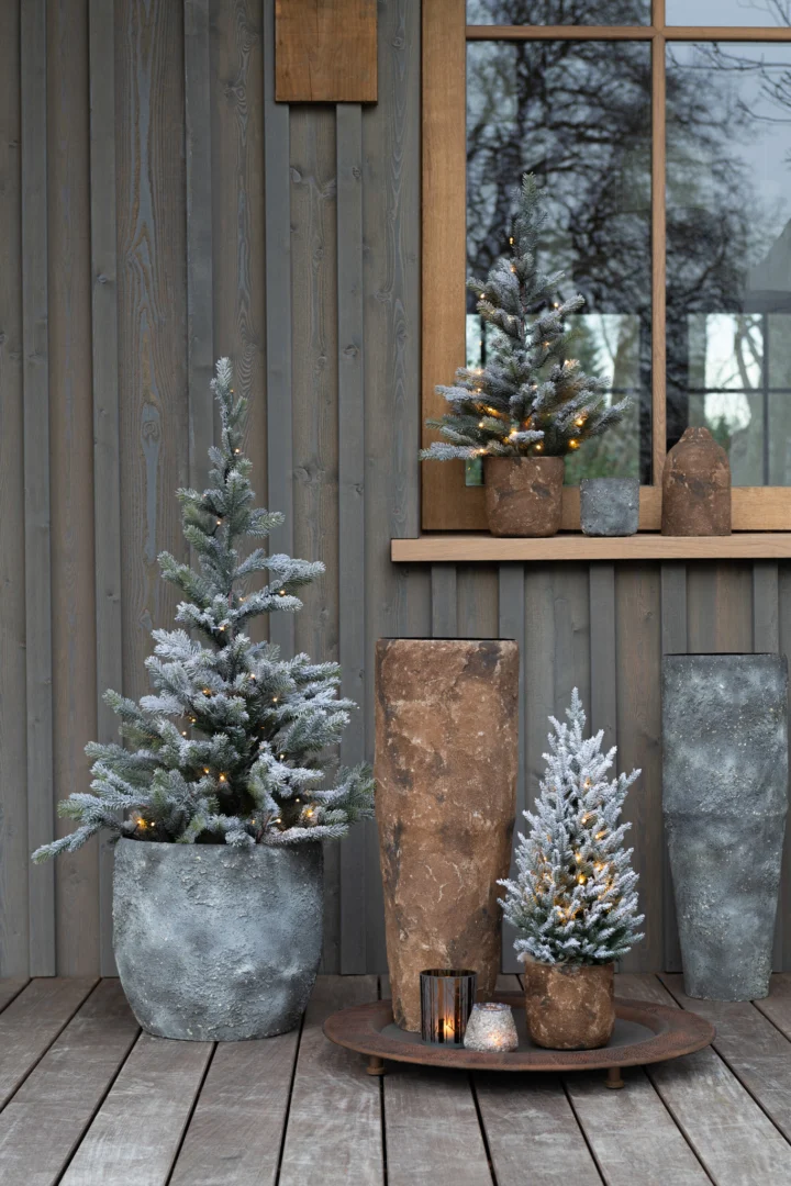 Drie kunstmatige besneeuwde dennenbomen in pot met koordverlichting staan uitgestald op een houten veranda naast twee Kaarsenhouder Orkaan Boom Glas Bruin Medium kandelaars, voor een grijze muur en raam die kale wintertakken weerspiegelen.