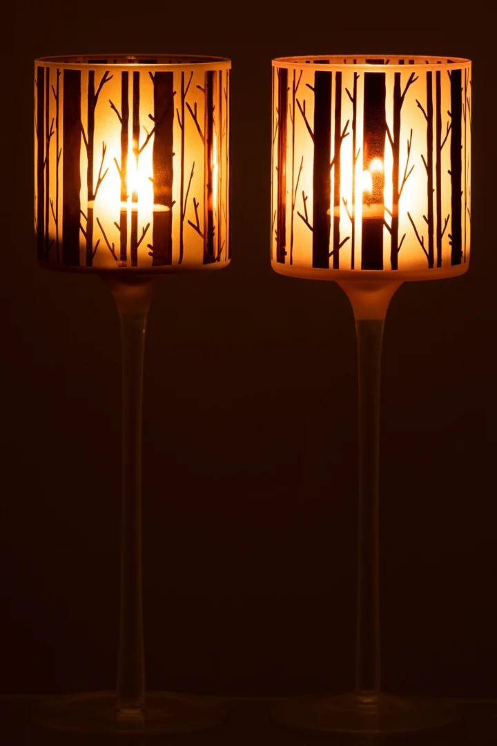Theelichthouder Op Voet Boom Glas Bruin Groot Set 2: Twee glazen kandelaars met boommotieven werpen warm licht en boomvormige schaduwen op hun kappen, die prachtig afsteken tegen een donkere achtergrond.