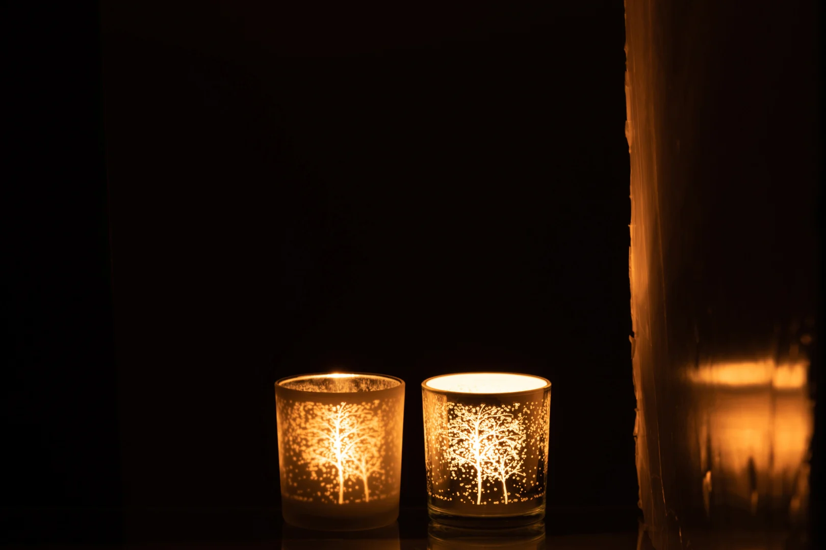 De Theelichthouder Boom Glas Wit Zilver Klein Set 2 heeft twee glazen kandelaars met boommotieven, die warm oplichten in een donkere kamer als kaarslicht elke houder zacht verlicht en reflecteert op een nabijgelegen oppervlak.
