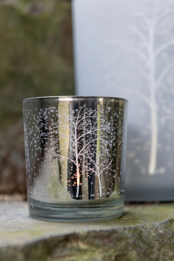 De Theelichthouder Boom Glas Wit Zilver Klein Set 2 heeft een zilverkleurig glas met een delicaat wit boom- en stippenpatroon, weergegeven op steen, met een soortgelijk ontwerp zacht vervaagd op de achtergrond.