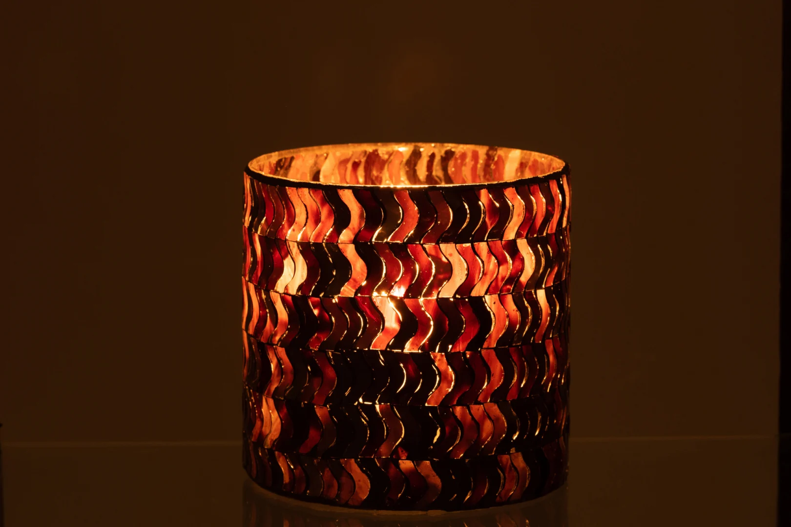 De Kaarsenhouder Orkaan Golf Glas Donkerroze heeft een brandende kaars in een cilindrische houder met golvende patronen in donkerroze tinten, die een warm, gloeiend licht werpt in het donker.