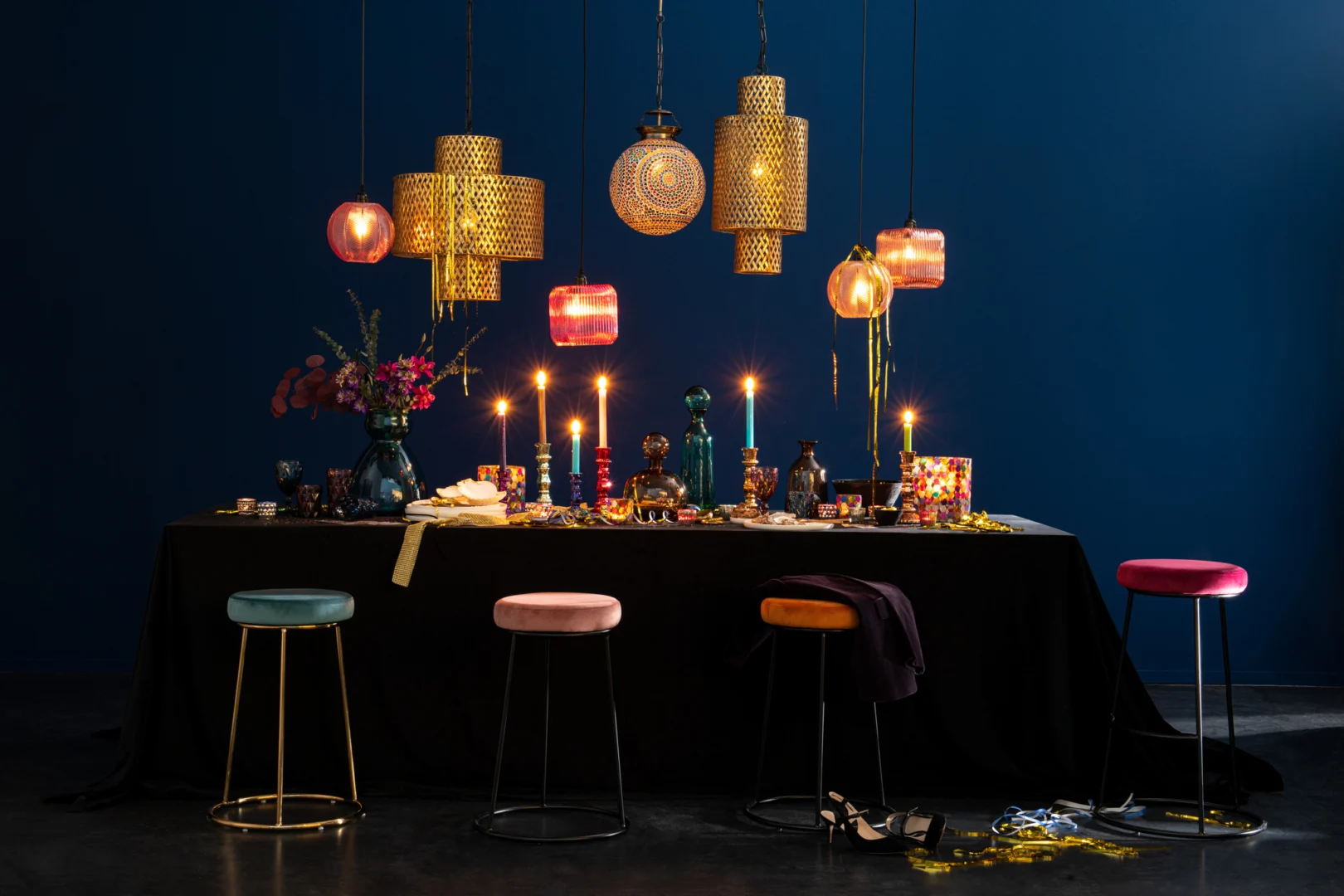 Een donkere, elegante eettafel gedekt voor een maaltijd met fluwelen krukjes en bloemen gloeit bij kaarslicht, met de Kaarsenhouder Hurricane Glas Pub Mix Medium. Kleurrijk decor voegt charme toe als schoenen en linten verspreid op de vloer liggen onder hangende lampen.