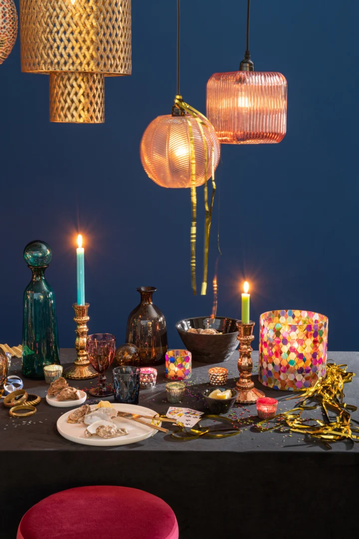 Op een feestelijke tafel staat de Kaarsenhouder Hurricane Glas Pub Mix Medium temidden van kleurrijk glaswerk, kaarsen en decoraties; hanglampen en gouden slingers erboven, met een blauwe muur en een rood kussentje ervoor.