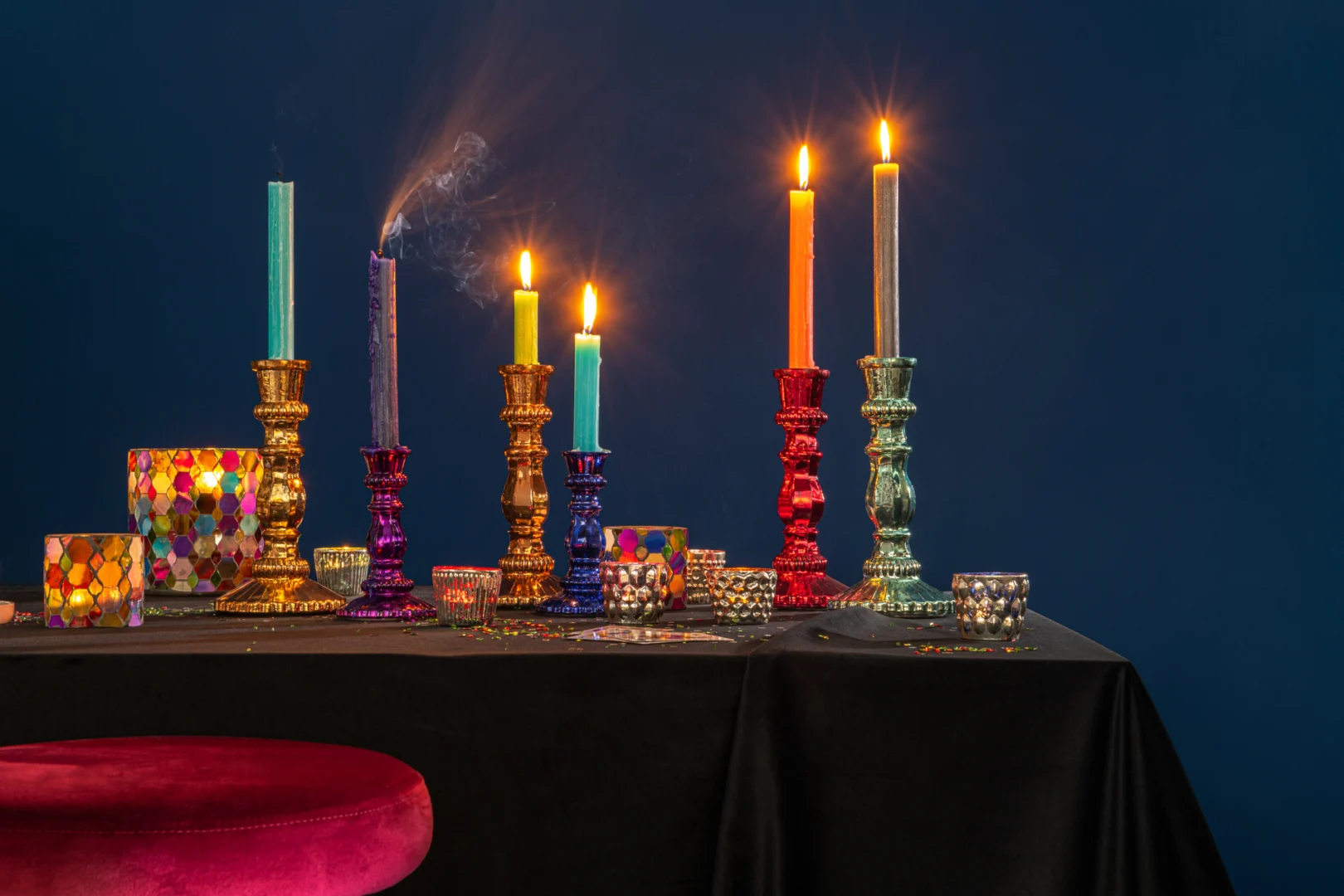Op een zwarte tafel staat de Kaarsenhouder Hurricane Glas Pub Mix Medium met vijf hoge, brandende kaarsen in veelkleurige houders; rook krult uit een net gedoofde kaars. Een rood fluwelen krukje staat ervoor, alles tegen een diepblauwe achtergrond.