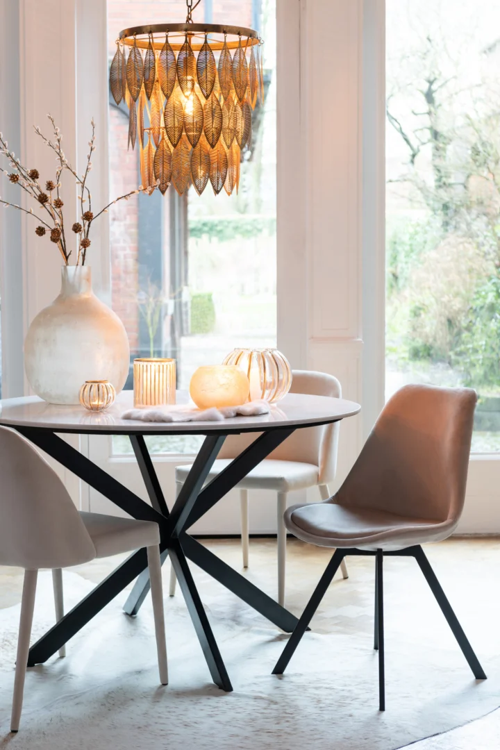 Een moderne eethoek met een ronde tafel, vier beige stoelen, de Kaarsenhouder Orkaan Bal Streep Glas Goud Klein, decoratieve vazen en een hanglamp in de vorm van een gouden blad bij grote ramen met uitzicht op een tuin.