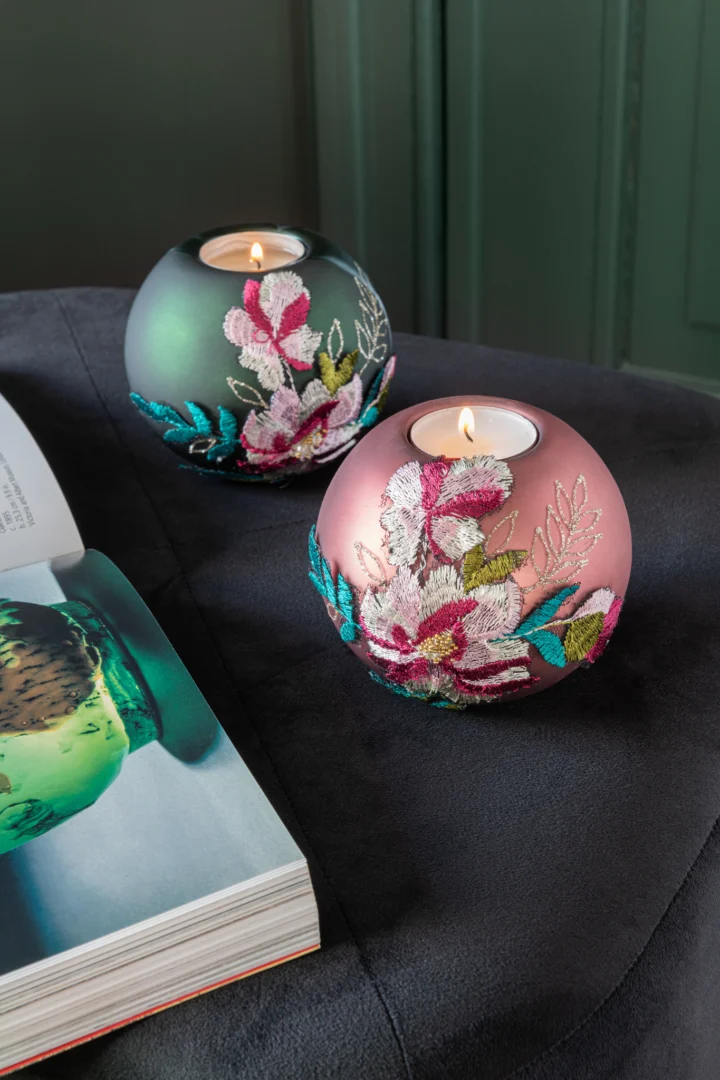 Twee ronde Theelichthouders Geborduurd Bloemglas Bordeaux kandelaars, versierd met kleurrijk bloemenborduurwerk, staan op een donker oppervlak bij een open tijdschrift. Beide houden een brandend waxinelichtje binnenin. Set van 4.