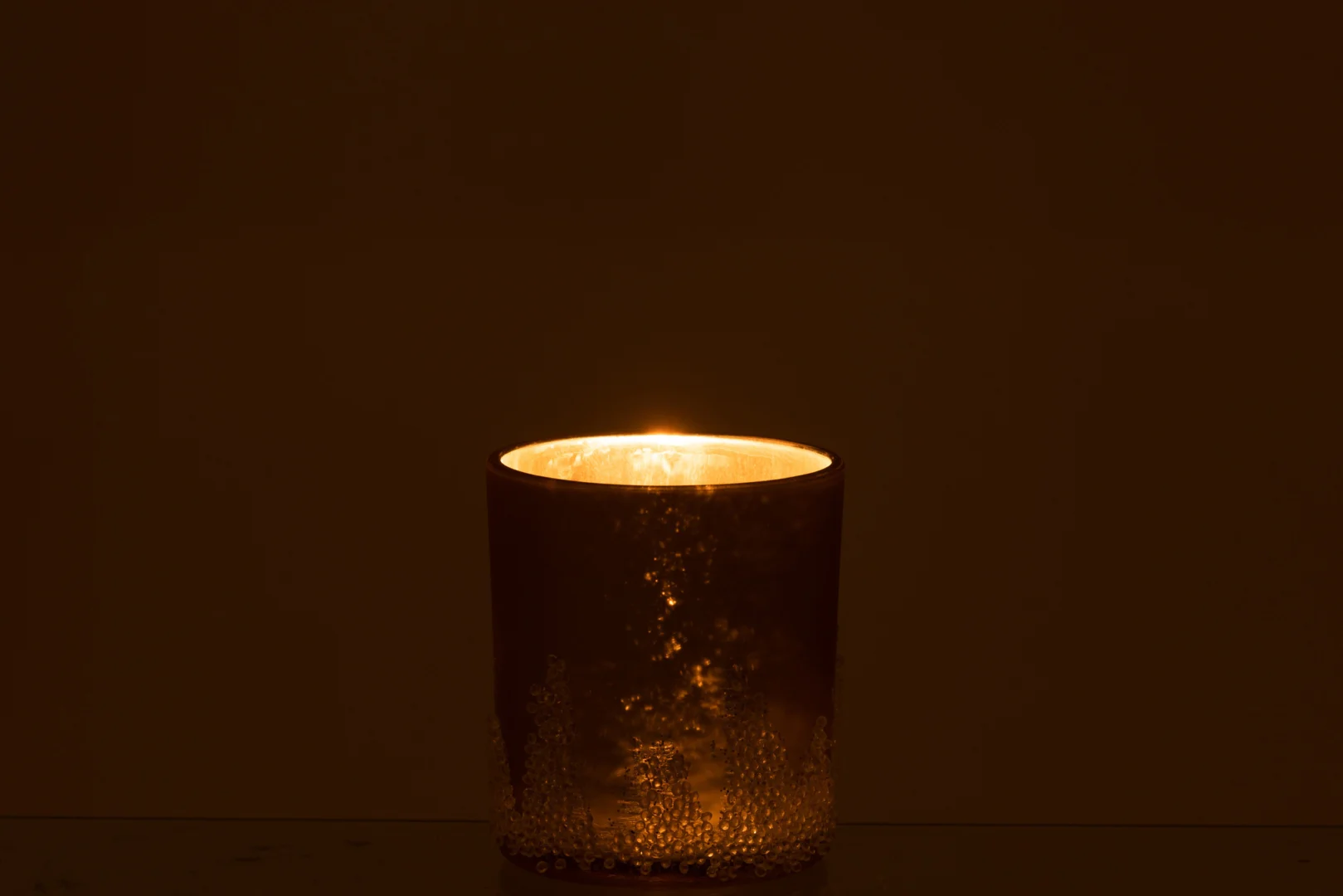 Een enkele brandende kaars gloeit warm in het donker, geplaatst in de Theelichthouder Suikerglazuur Glas Goud Groot - een matglazen houder met delicate structuurpatronen.