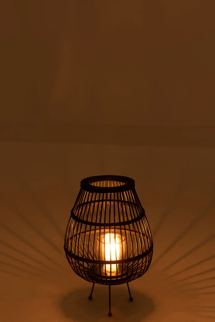 Een brandende kaars in de Lantaarn Daya Hout Zwart Klein straalt een warme gloed en schaduwen uit op een effen, bruin oppervlak.