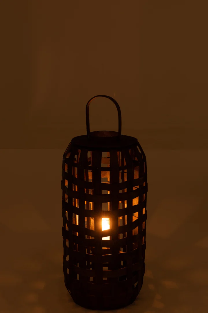 De Lantaarn Cilinder Hout Donkerbruin Klein is een kleine, gevlochten houten lantaarn met een handvat dat warm oplicht in het donker en zacht licht en schaduwen werpt door het rasterontwerp.