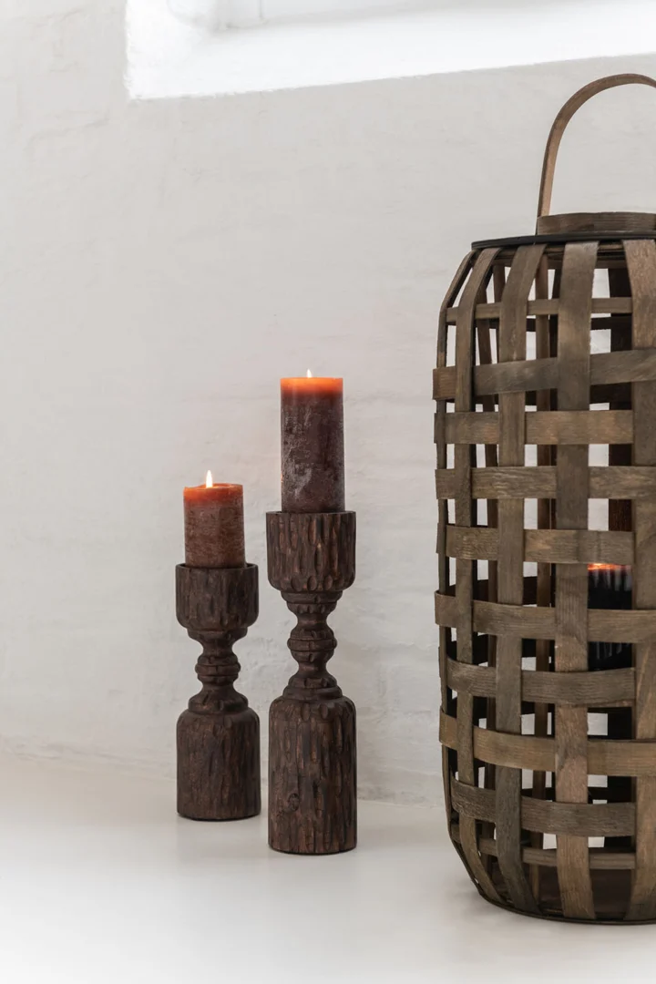 Een Lantaarn Cilinder Hout Donkerbruin Klein staat naast een grote gevlochten houten mand met handvat, beide op een wit vlak tegen een witte muur.