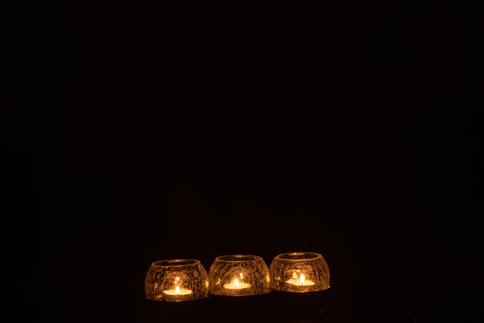 Drie Theelichthouder Bal Craquelure Glas RVS Zilver kandelaars, elk met een brandend waxinelichtje, staan naast elkaar tegen een donkere achtergrond, waardoor een warme en rustige zilveren gloed ontstaat.