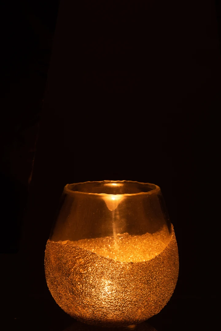 De Theelichthouder Halfparels Ronde Glas Goud Medium gloeit warm in het donker, werpt een zacht, gouden licht en creëert een gezellige, sfeervolle sfeer tegen een zwarte achtergrond.