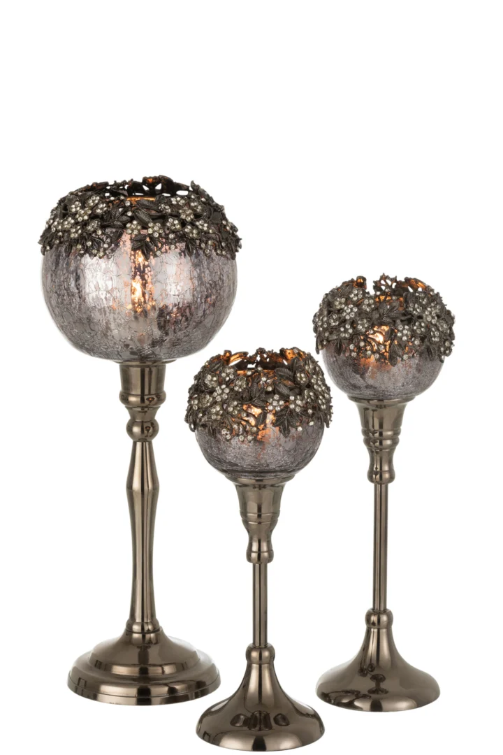 De Theelichthouder Op Voet Juweel Glas Smokey Grijs Groot is een metalen kandelaar met een rond, gestructureerd rookgrijs glazen blad en decoratieve bloemenaccenten op de rand, afgebeeld met een brandende kaars tegen een witte achtergrond.