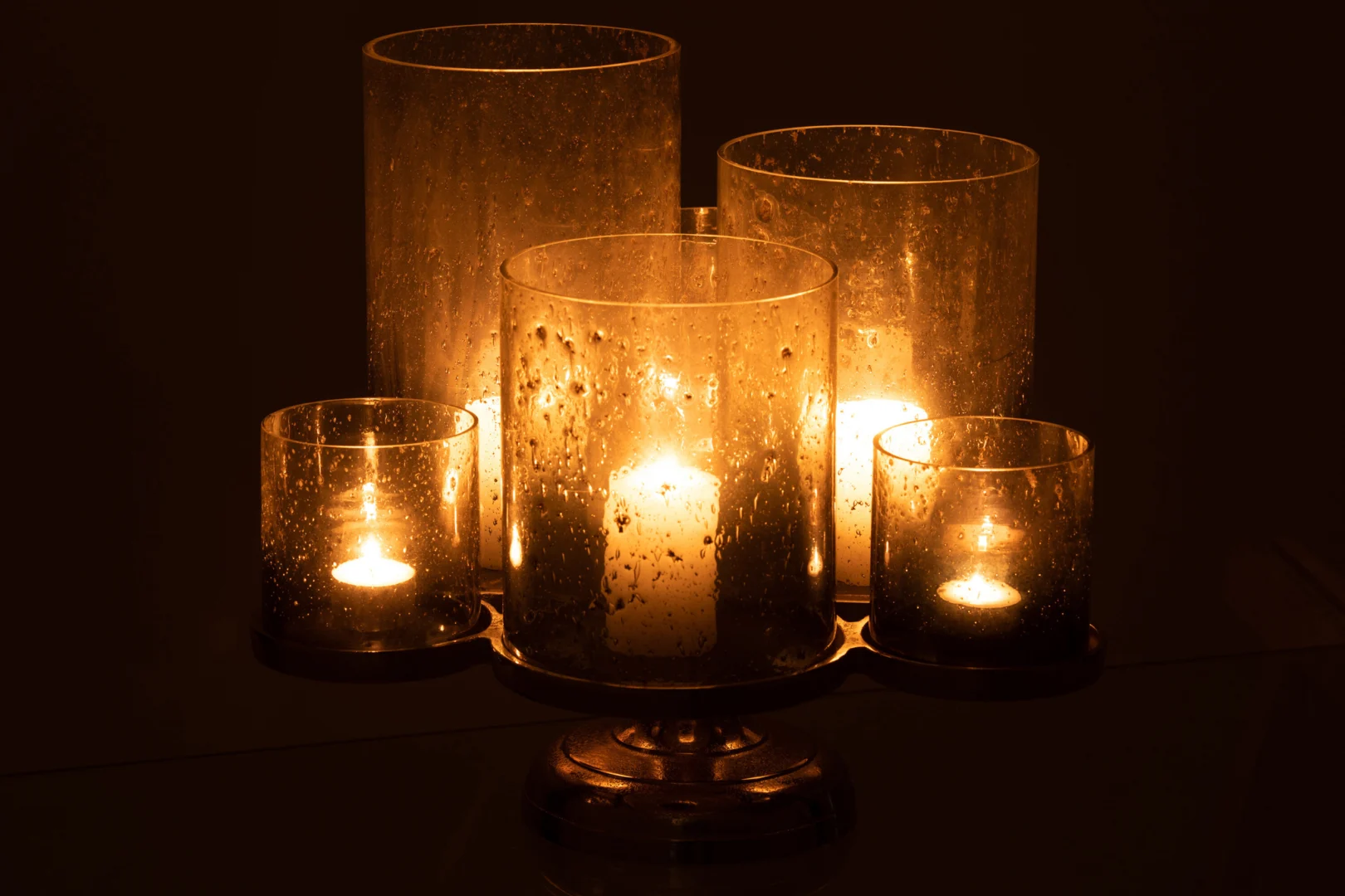 Vijf Kaarsenhouder Orkaan Op Voet Glas Zilver Grijs in verschillende hoogtes gloeien warm op in het donker, hun gespikkelde glazen houders werpen een gestructureerd gouden licht.