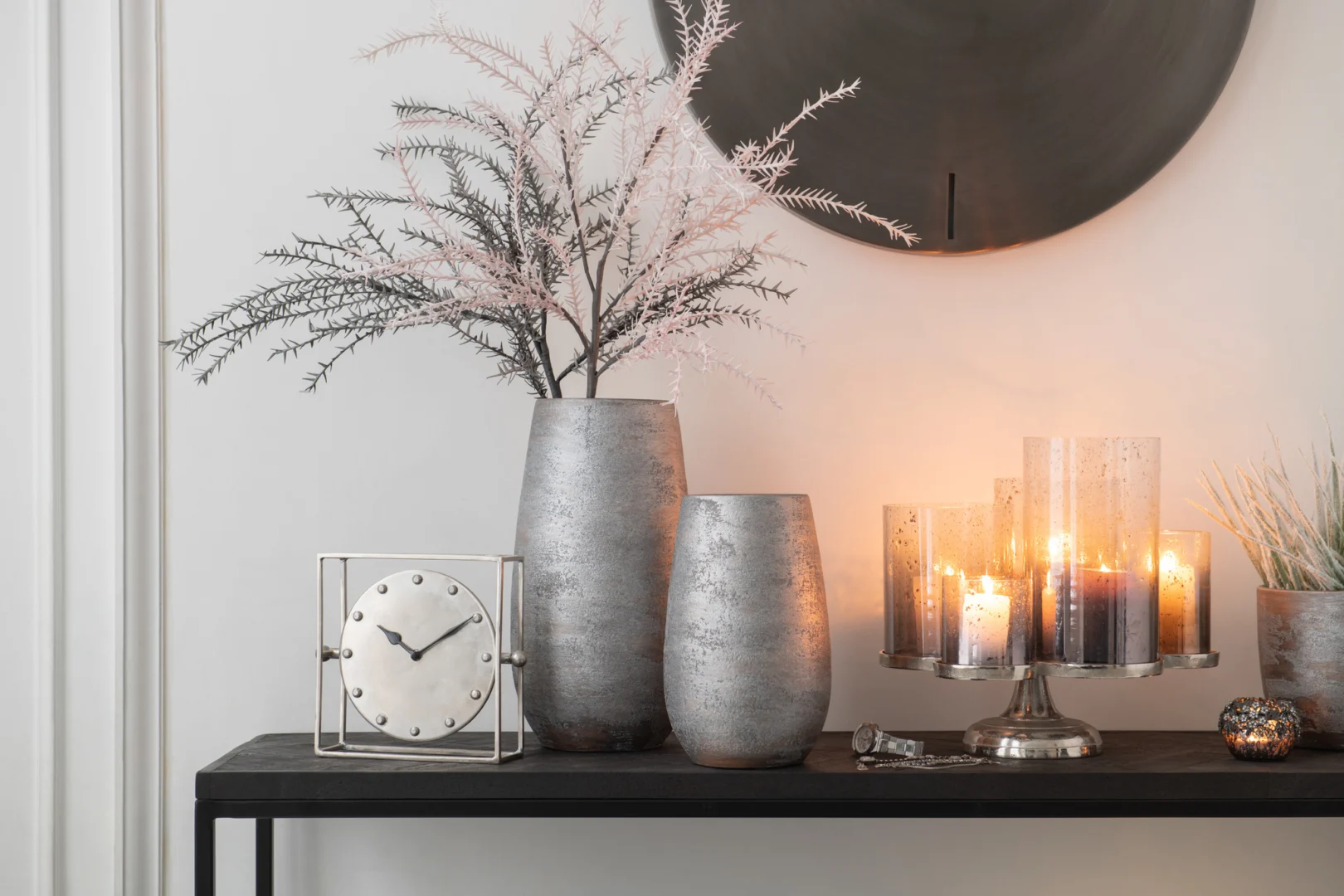 Een moderne consoletafel is voorzien van Kaarsenhouder Orkaan Op Voet Glas Zilver Grijs kandelaars, zilveren vazen, een zilveren klok en decoratieve takken. Een ronde spiegel erboven maakt de elegante en gezellige setting compleet.