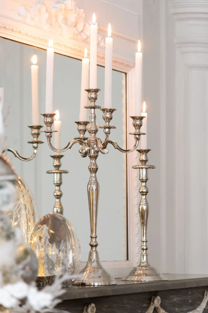 De Kaarsenhouder Klassiek Aluminium Zilver Groot met brandende witte kaarsen staat op een schouw naast twee bijpassende zilveren houders, dit alles weerspiegeld in een decoratieve spiegel in een elegant klassiek interieur.
