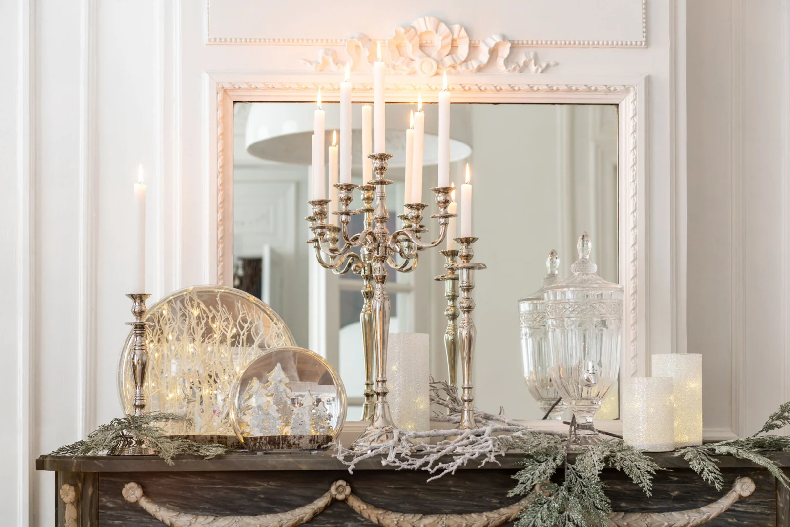 Een schouw versierd met de Kaarsenhouder Klassiek Aluminium Zilver Groot met brandende kaarsen, glazen potten, een sneeuwbol, een glazen stolp met winterlandschap en feestelijk groen staat voor een ingelijste spiegel.