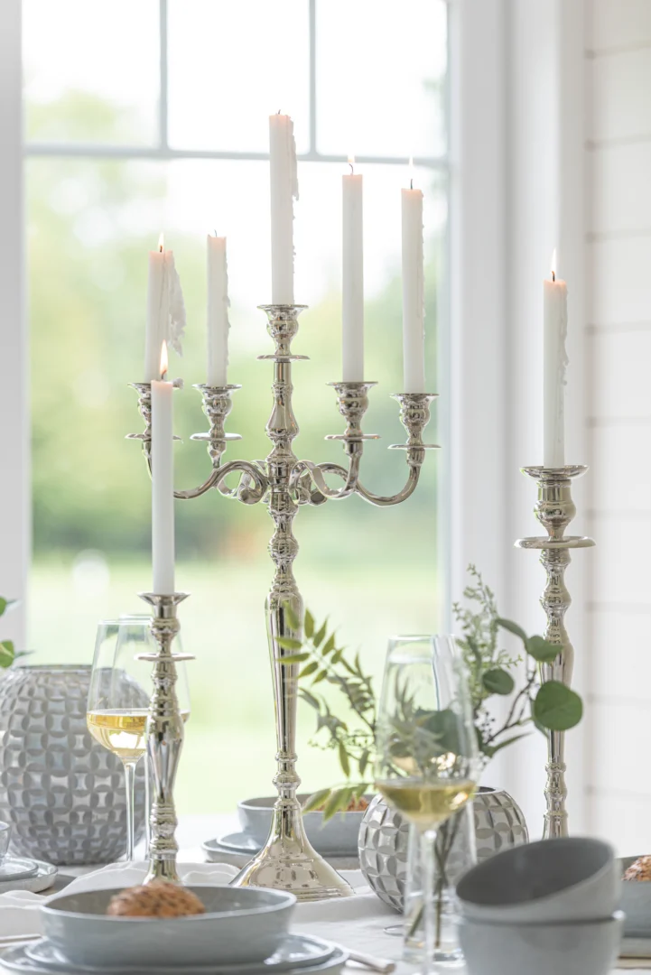 Een heldere, elegante tafelschikking met de Kaarsenhouder Klassiek Aluminium Zilver Groot met hoge witte kaarsen, wijnglazen, borden met eten, decoratieve vazen en groen gebladerte bij een groot raam met natuurlijk licht.