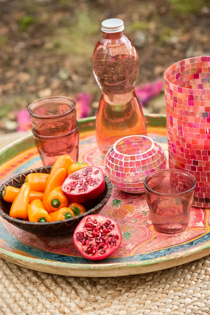 Op een kleurrijk dienblad staan twee glazen, een schaaltje oranje minipaprika's, een gehalveerde granaatappel, een waterflesje en Theelichthouder Cilinder Mozaïek Glas Roze Mix Groot op een geweven mat buiten.