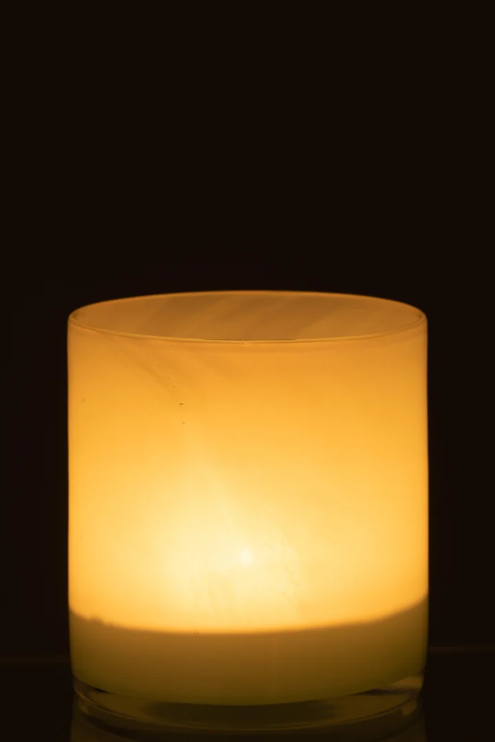 De Theelichthouder Jade Glas Lichtgroen Medium gloeit in het donker en werpt een zacht geel licht dat het gladde jadegroene glas zachtjes accentueert, terwijl de achtergrond volledig zwart blijft.