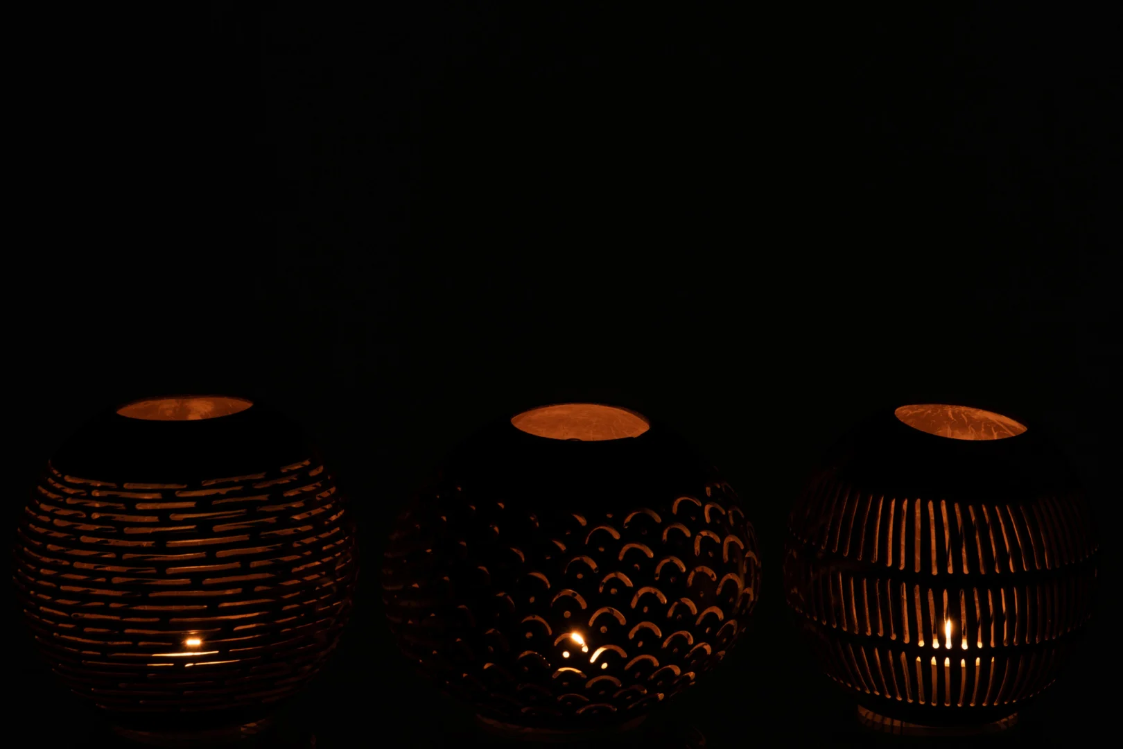 De Theelichthouder Kokosnootlijnen Bruin Assortiment Van 3 heeft ronde kandelaars met gedetailleerde uitsnijdingen die zachtjes gloeien en warm, gedessineerd licht werpen door hun ontwerpen van kokosnootschelpen tegen een donkere achtergrond.