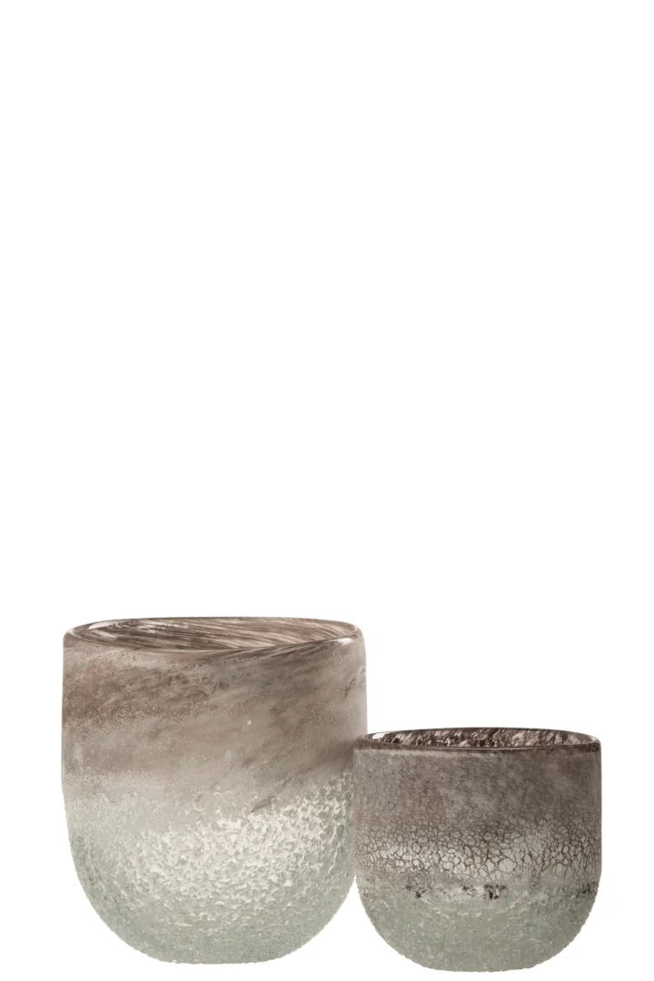 De Kaarsenhouder Orkaan Scavo Glas Grijs Groot is een ronde glazen vaas met een mat, gestructureerd oppervlak en een verloop van grijs aan de bovenkant naar helder aan de onderkant. Het grotere formaat is ontworpen om achter een kleinere versie te plaatsen.