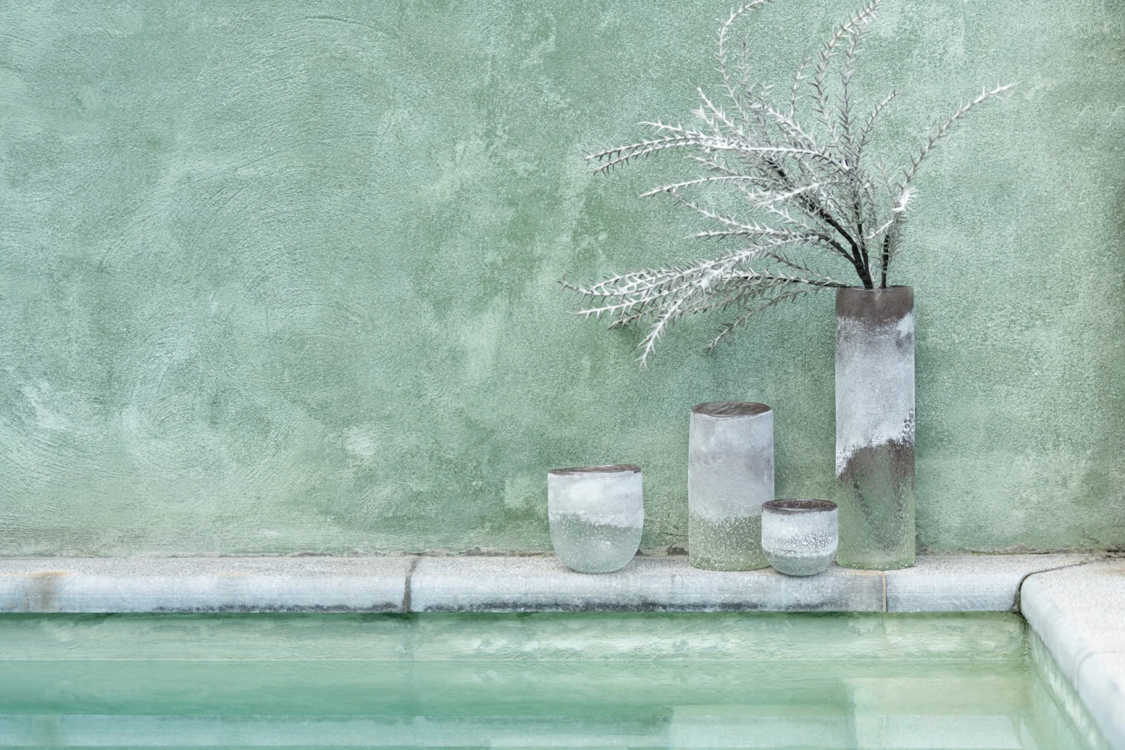 De Kaarsenhouder Orkaan Scavo Glas Grijs Groot staat bij een lichtgroen zwembad, het minimalistische ontwerp voegt een rustige, eigentijdse touch toe tegen een groene muur met textuur.