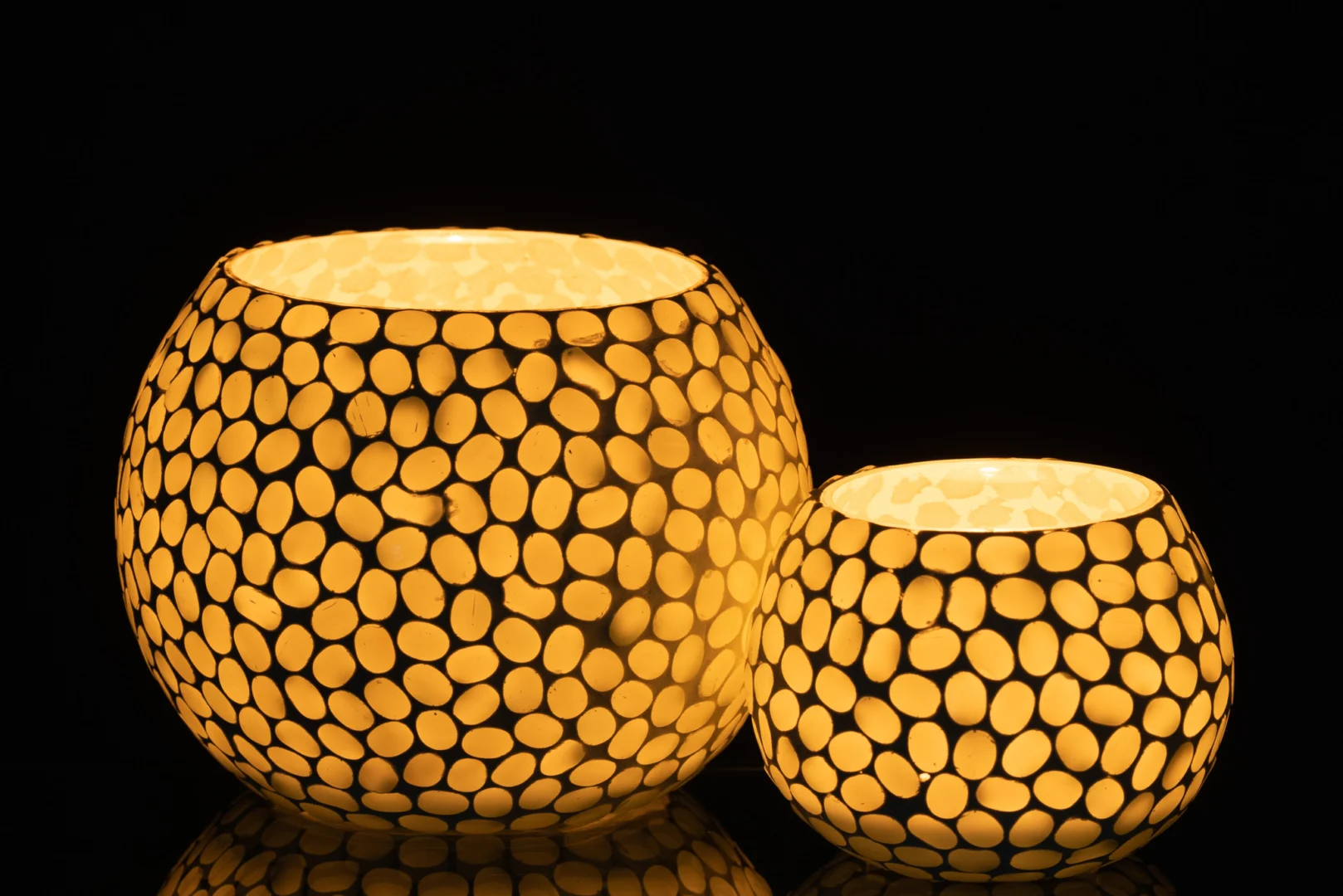 De Theelichthouder Orb Glas Wit Aqua Groot heeft een rond ontwerp met een ovaal mozaïekpatroon, dat een warme gloed werpt tegen een zwarte achtergrond voor een gezellige, uitnodigende sfeer.