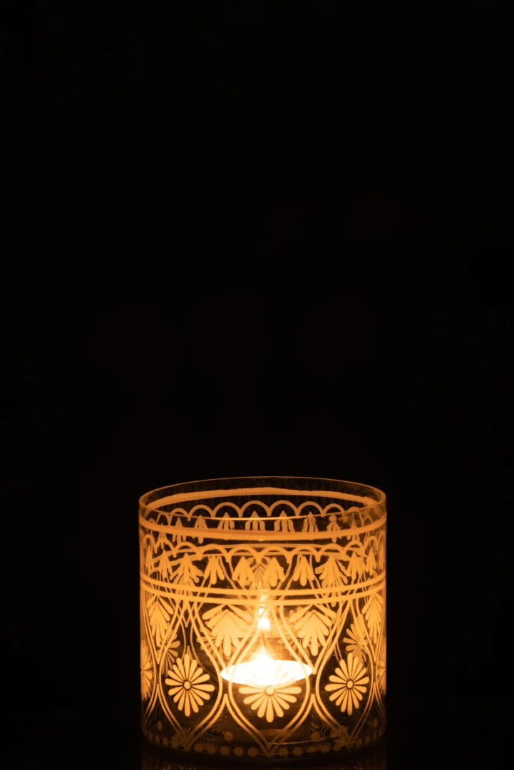 De Kaarsenhouder Orkaan Glas Transparant Wit Patroon Klein houdt een zacht gloeiende kaars vast, waarvan het decoratieve glazen ontwerp prachtig afsteekt tegen een donkere achtergrond.