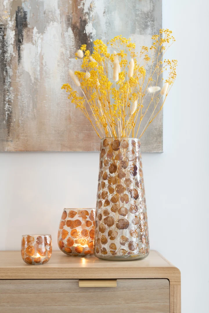 De Orkaan Gerard Glas Amber Groot vaas, met een bruin en wit gevlekt patroon, houdt gele droogbloemen op een houten kastje naast twee bijpassende kandelaars. Aan de muur erachter hangt een abstract schilderij.