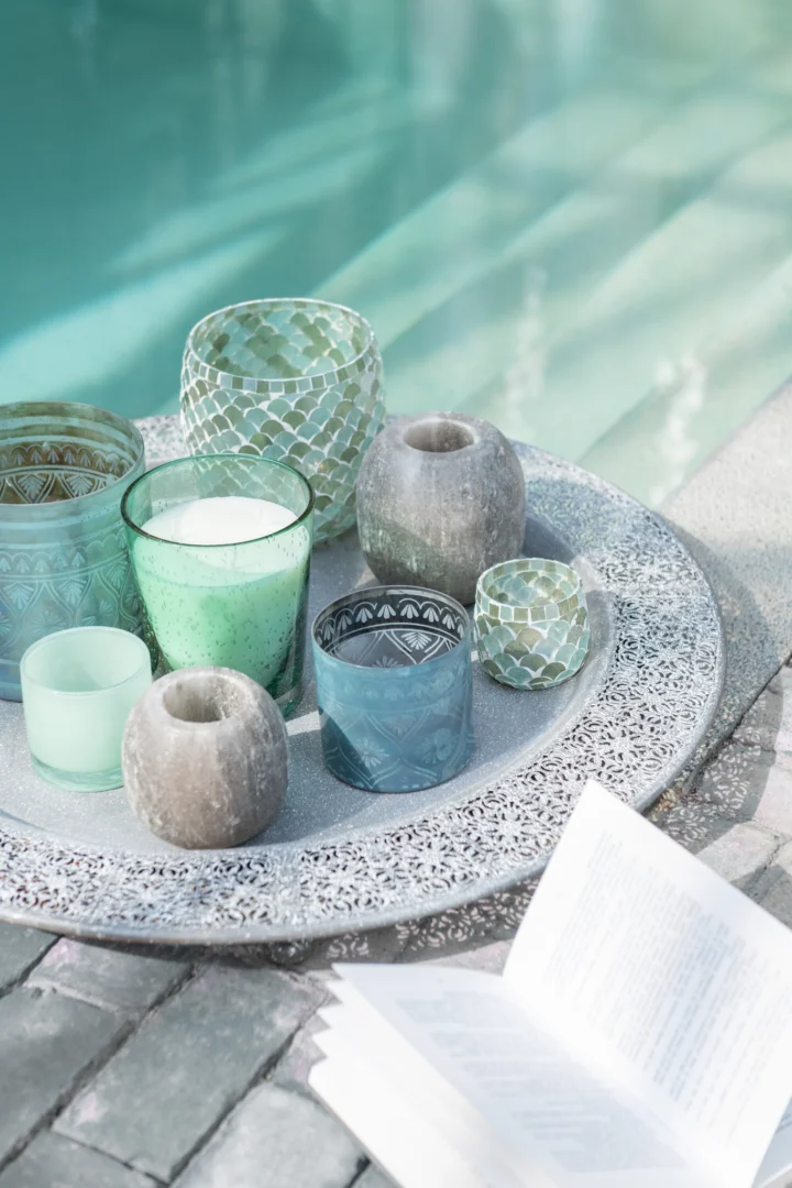 Het Kaarsenhouder Orkaan Mozaïekglas Aqua Medium siert het zwembad, in combinatie met een open boek op stenen tegels, wat een ontspannen en serene sfeer creëert.