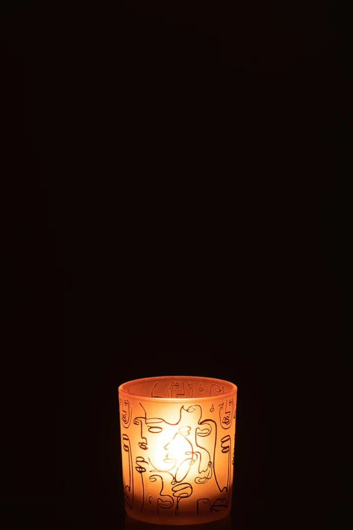Een enkele aangestoken Theelichthouder Happy Faces Glas Terracotta Klein gloeit zacht in zijn oranje houder met abstracte lijnmotieven tegen een volledig donkere achtergrond.
